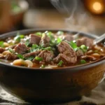 Steaming bowl of beef noodle soup with herbs and beef slices.